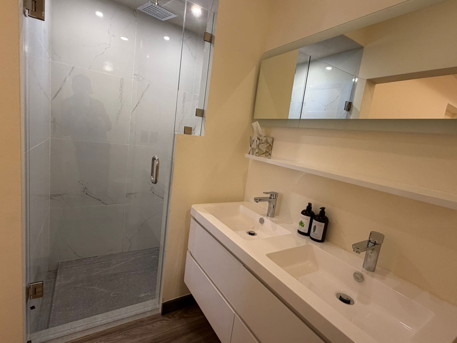 Un baño con cabina de ducha de vidrio, tocador blanco con dos lavamanos y un espejo sobre el tocador.