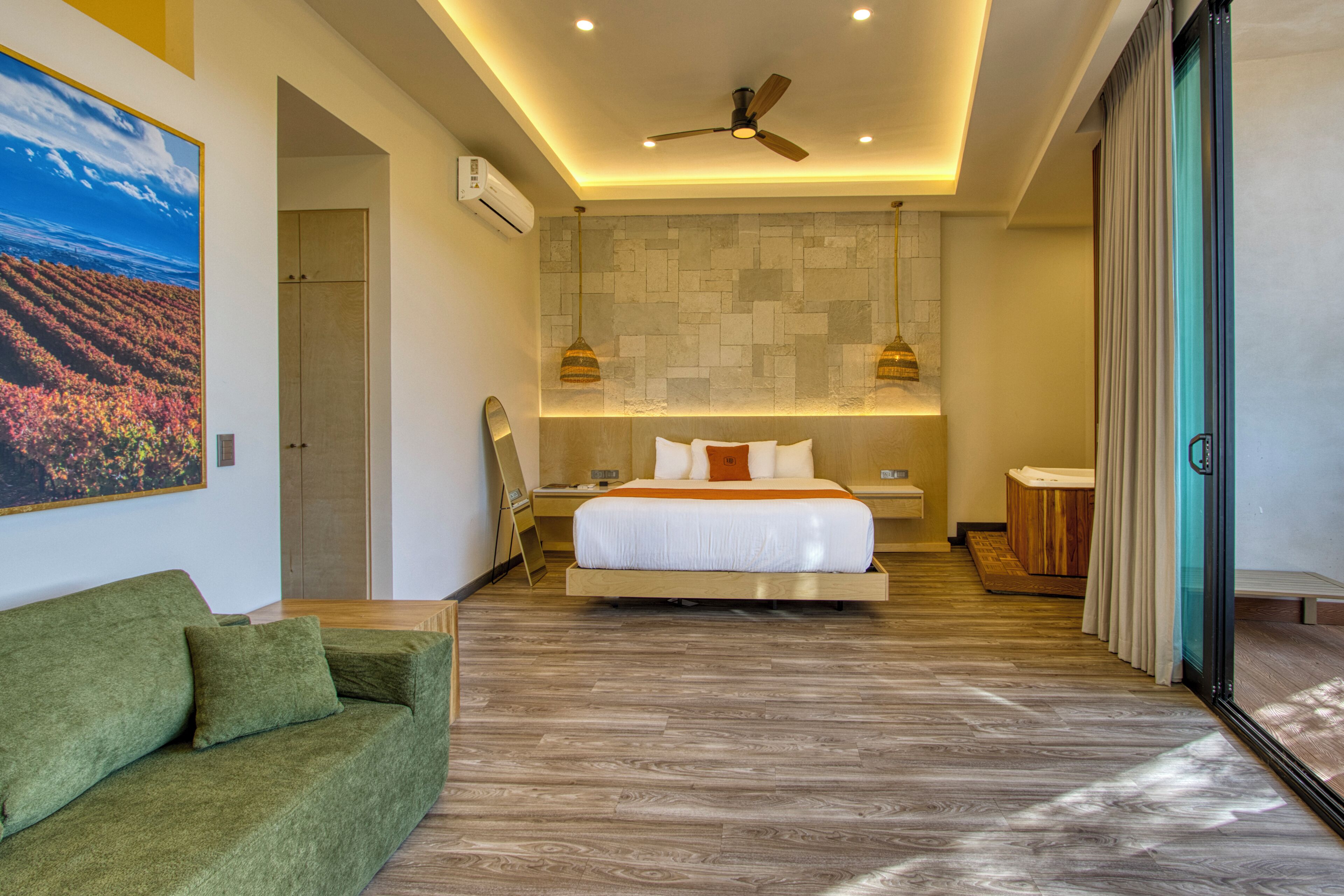 A modern hotel room with a bed, a green sofa, a ceiling fan, and a stone wall.