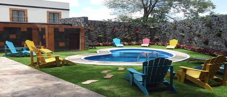 A backyard with a swimming pool, Adirondack chairs, a stone wall, and a grassy lawn.
