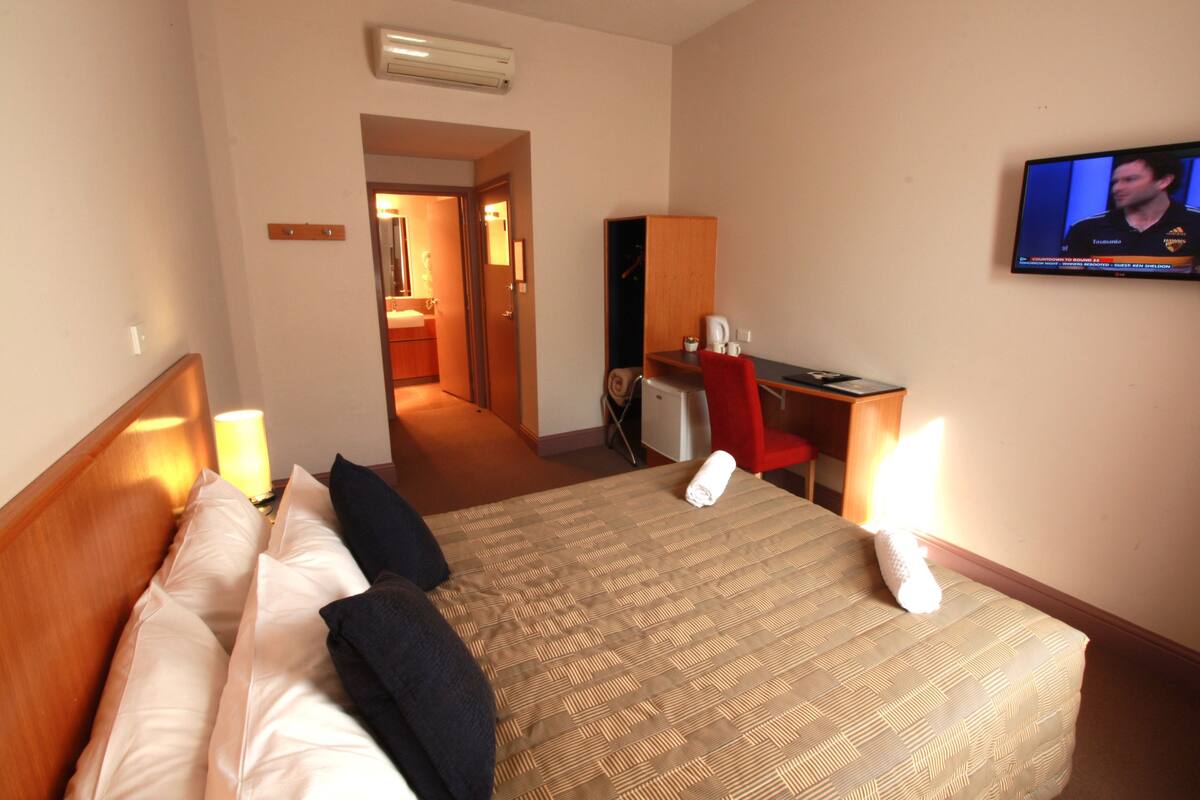 A hotel room with a bed, a desk, a chair, a television, and a bathroom visible through an open door.