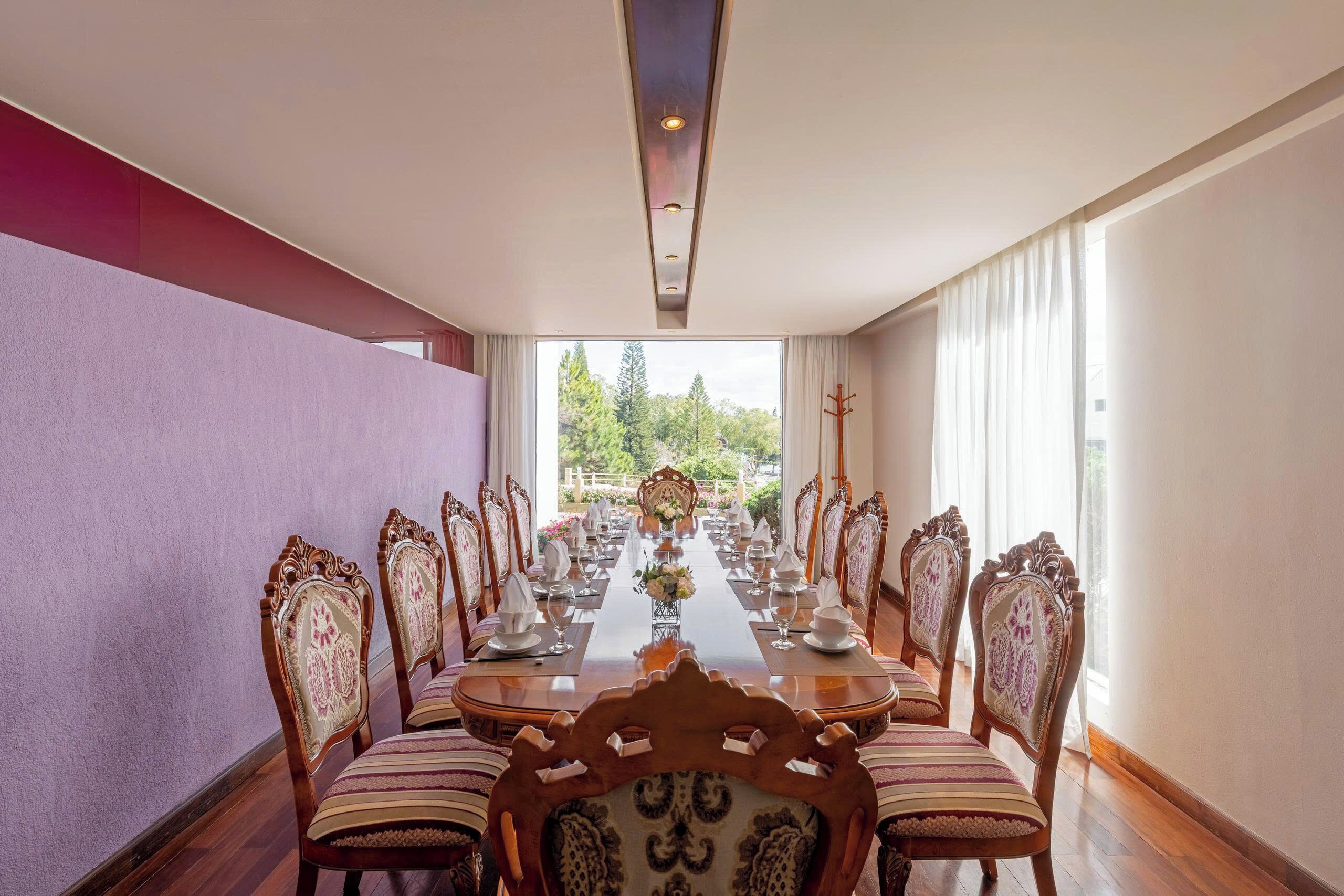 A dining room with a long table set for a meal, surrounded by ornate chairs, with a view of greenery outside.