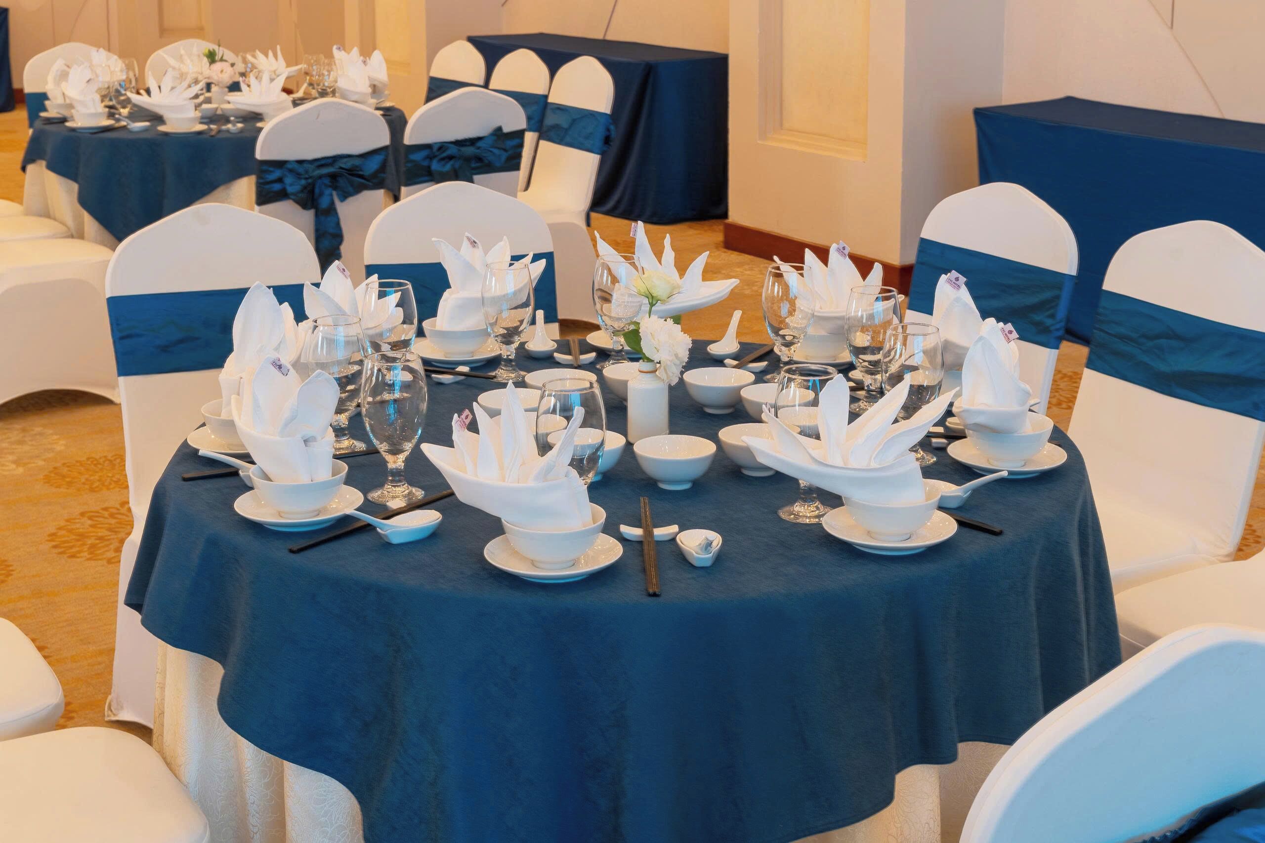 A banquet table set with white napkins, bowls, and glasses, surrounded by white chairs with blue sashes.
