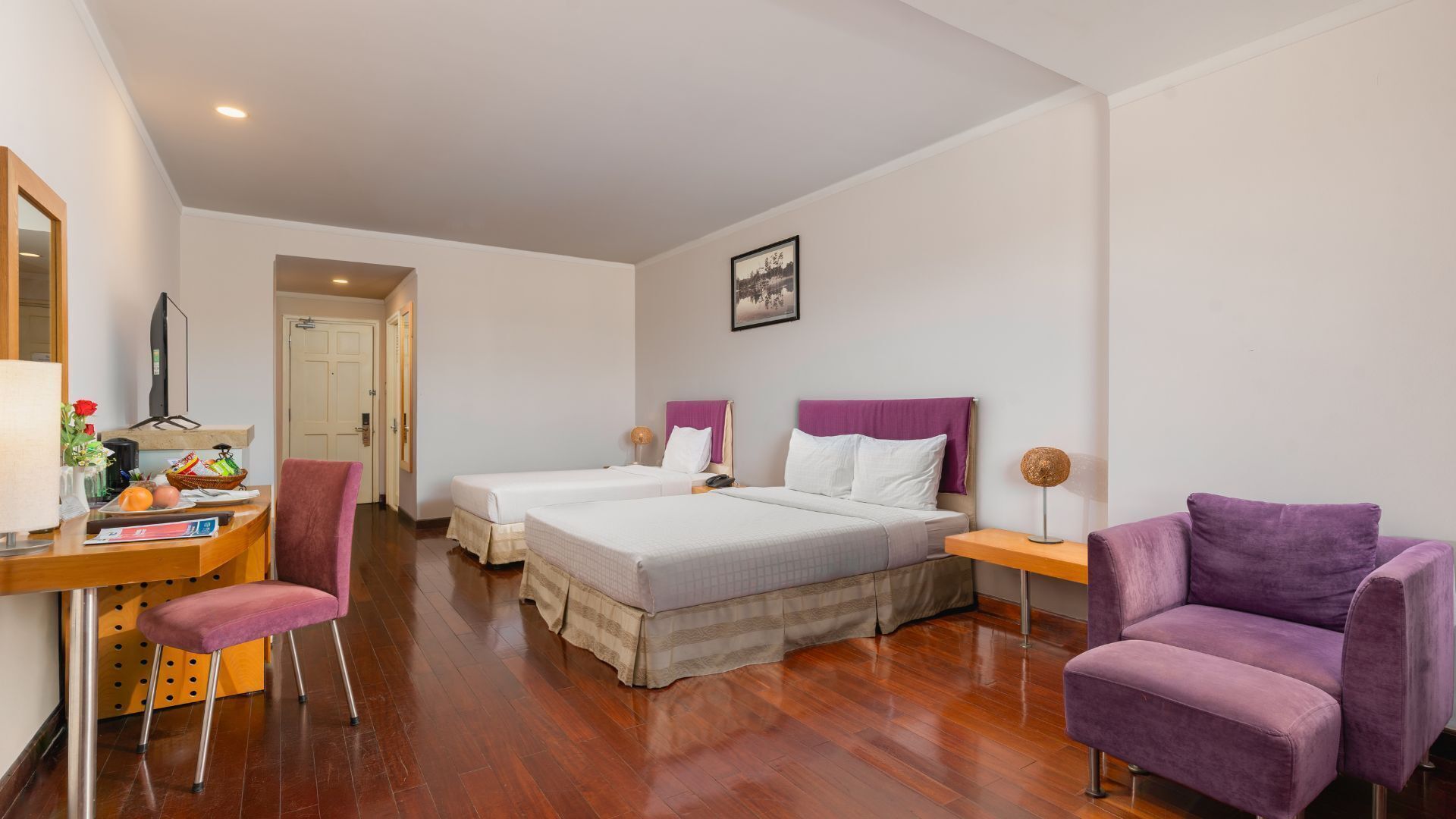 Une chambre d’hôtel avec deux lits, un bureau avec un ordinateur, une chaise et un fauteuil violet.