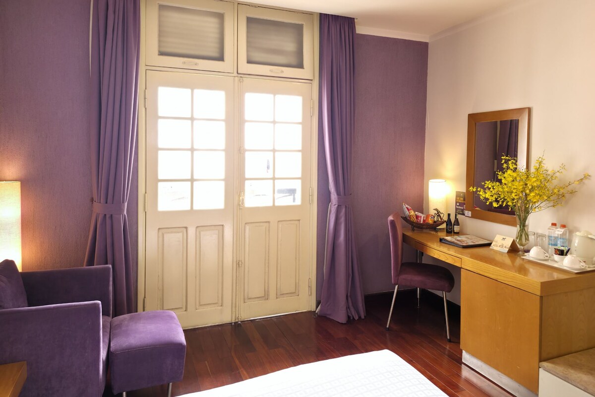 Habitación de hotel con escritorio de madera, sillas moradas, un ventanal con cortinas, un espejo y un florero con flores amarillas.
