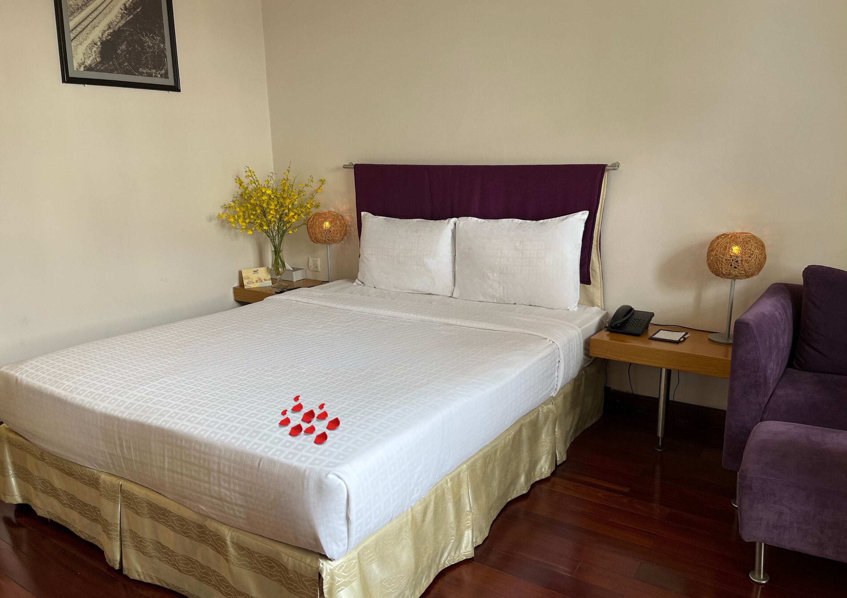 A neatly made bed with a white bedspread and a purple headboard, a bedside table with a lamp, a chair, and a small vase with yellow flowers.
