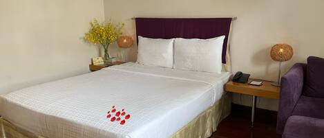 A neatly made bed with a white bedspread and a purple headboard, a bedside table with a lamp, a chair, and a small vase with yellow flowers.