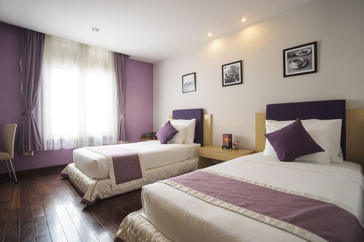 Ein Hotelzimmer mit zwei Betten, einem Sessel, einem Nachttisch und einem Fenster mit Vorhängen.