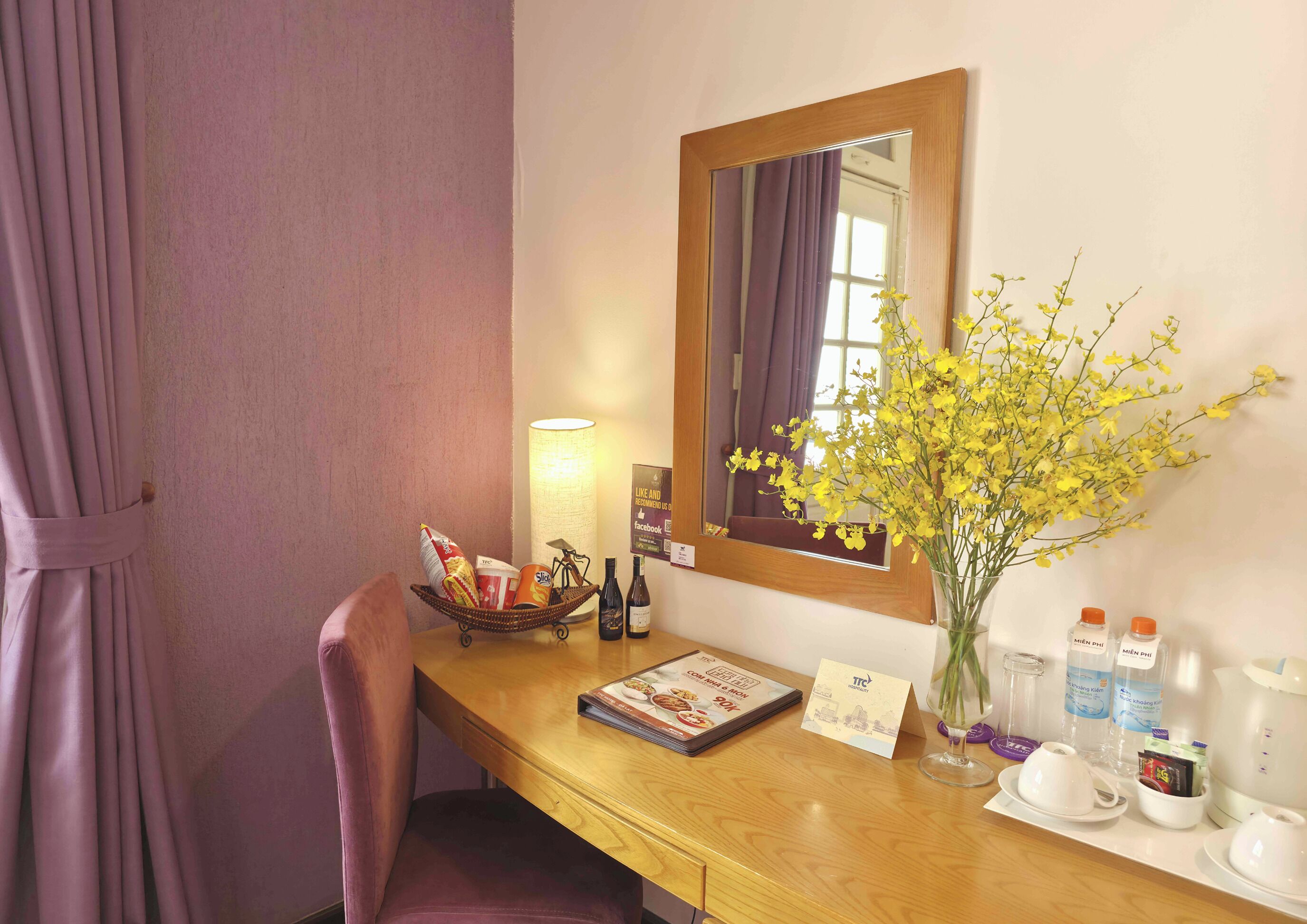 Una habitación de hotel con un escritorio de madera, un espejo, un jarrón con flores amarillas, una lámpara, un menú y un cartel con el nombre del hotel.