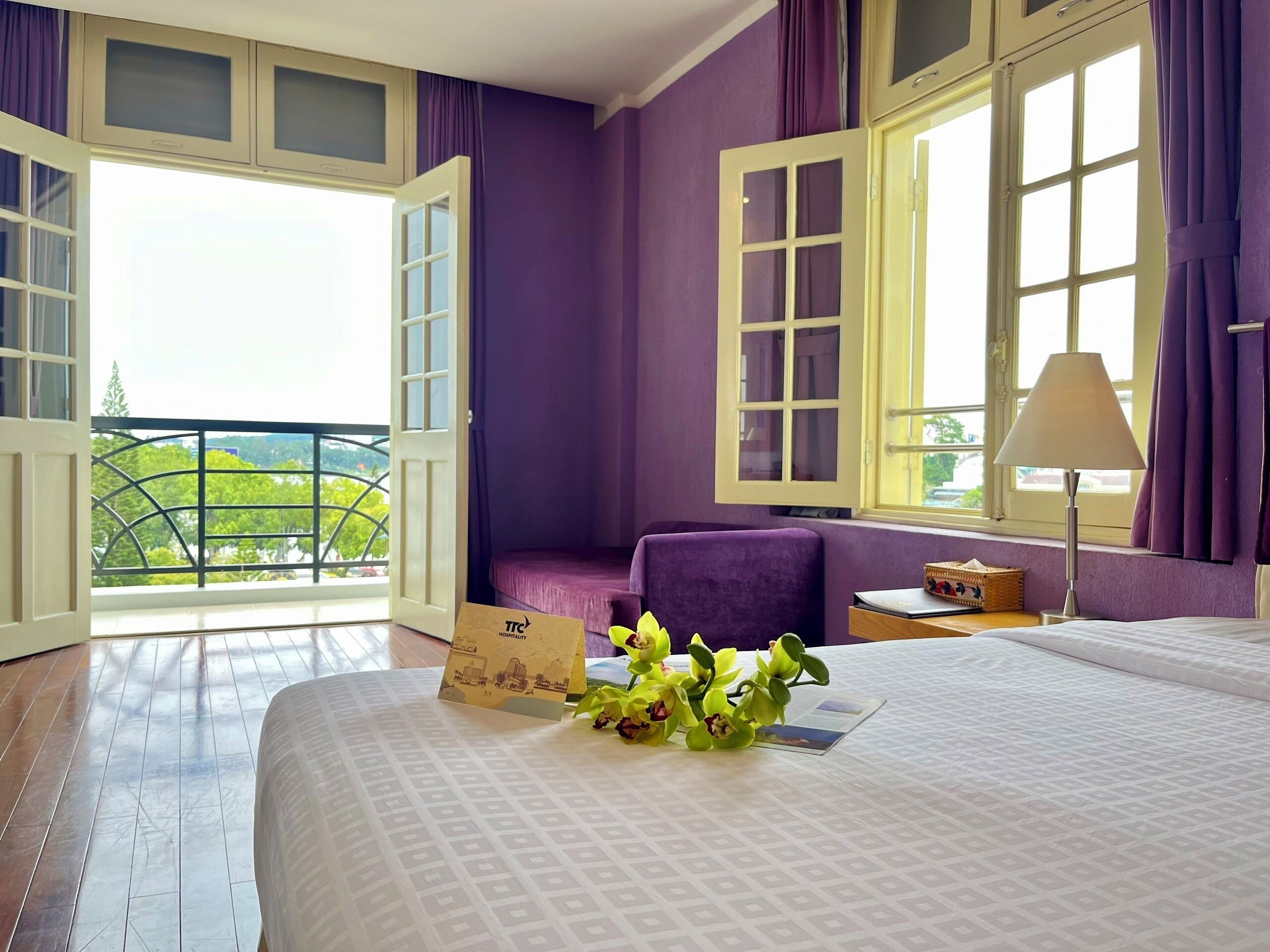 A hotel room with a balcony, a bed with a patterned bedspread, a small table with a lamp, and a chair.