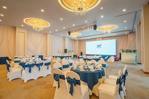 Een feestzaal met ronde tafels gedekt voor een formeel evenement, met witte stoelen met blauwe linten, kroonluchters en een projectiescherm waarop een logo staat.