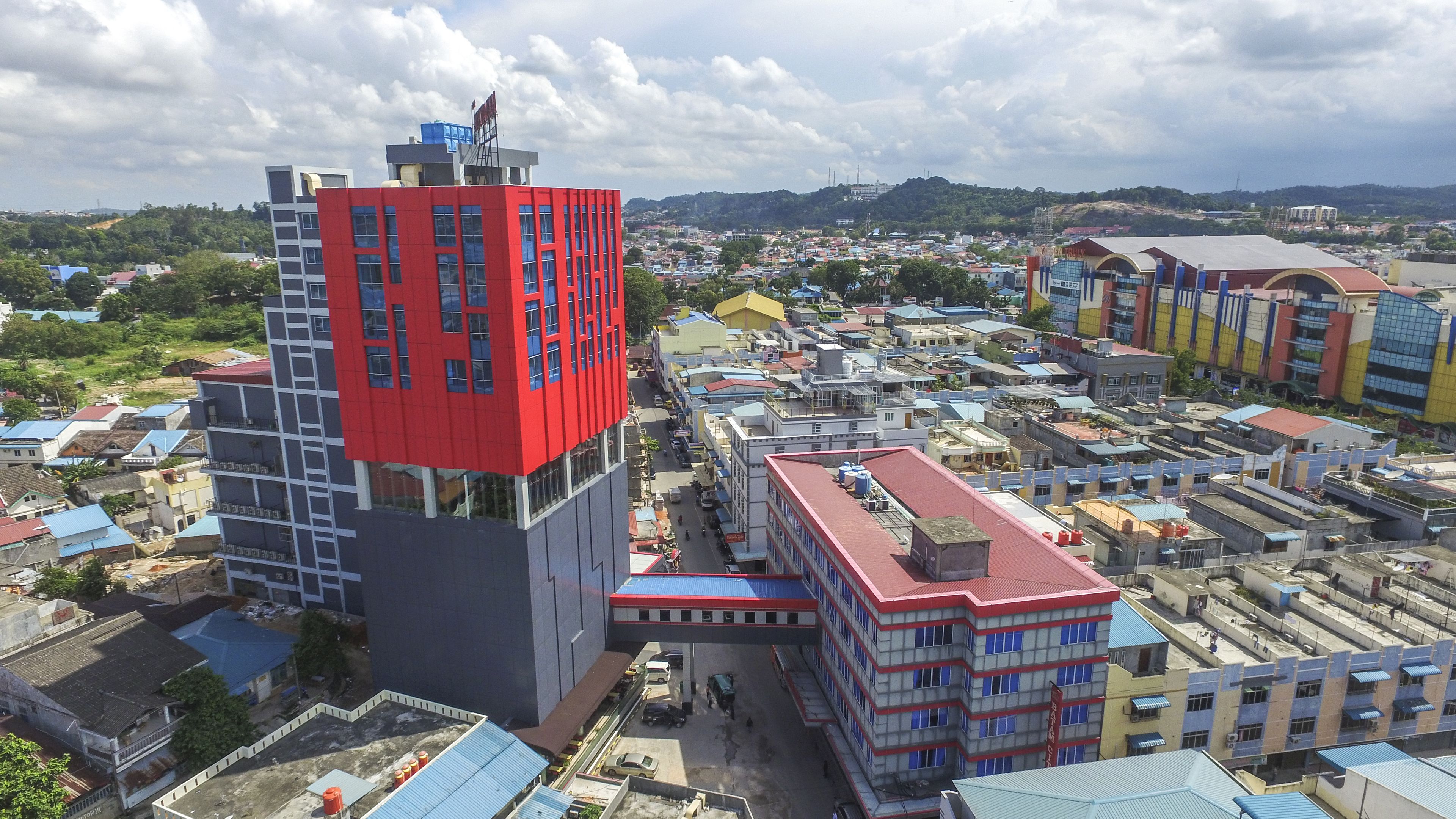 A modern building with a red and grey facade, surrounded by a densely packed urban area with various buildings and a hilly landscape in the background.