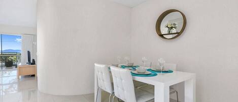A dining area with a white table set for four, a round mirror on the wall, and a view of the outdoors through a glass door.