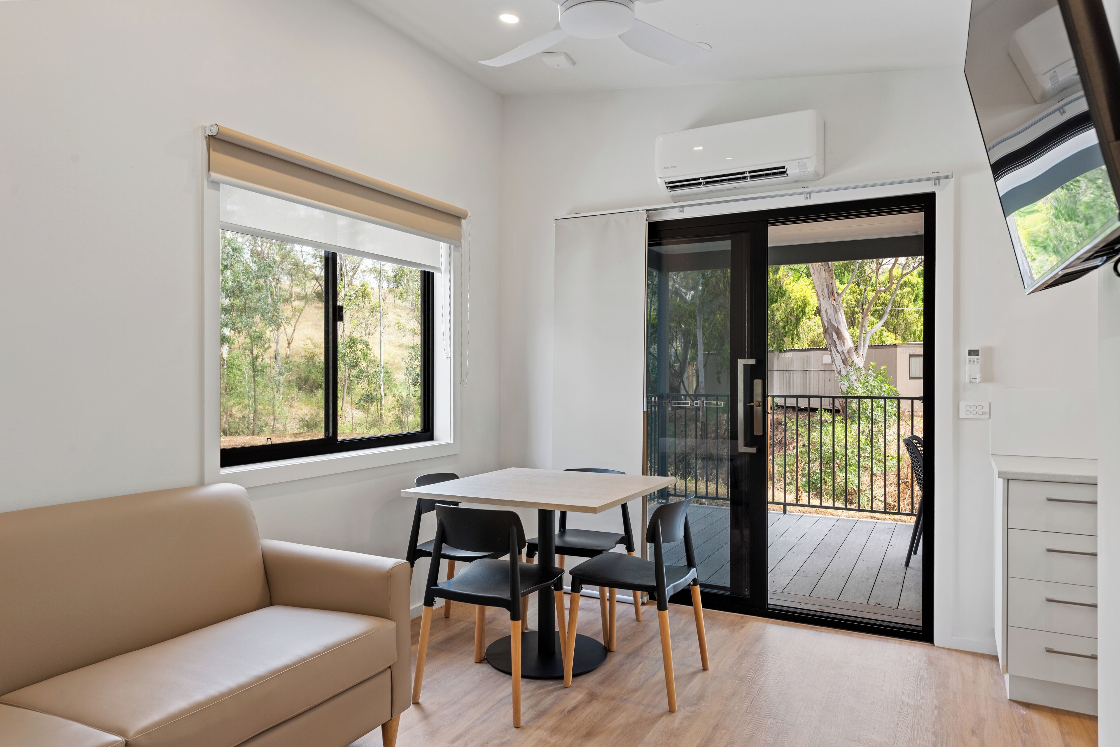 Una habitación con un sofá color beige, una mesa con cuatro sillas, un ventilador de techo, un equipo de aire acondicionado y un ventanal corredero que da a una terraza al aire libre.