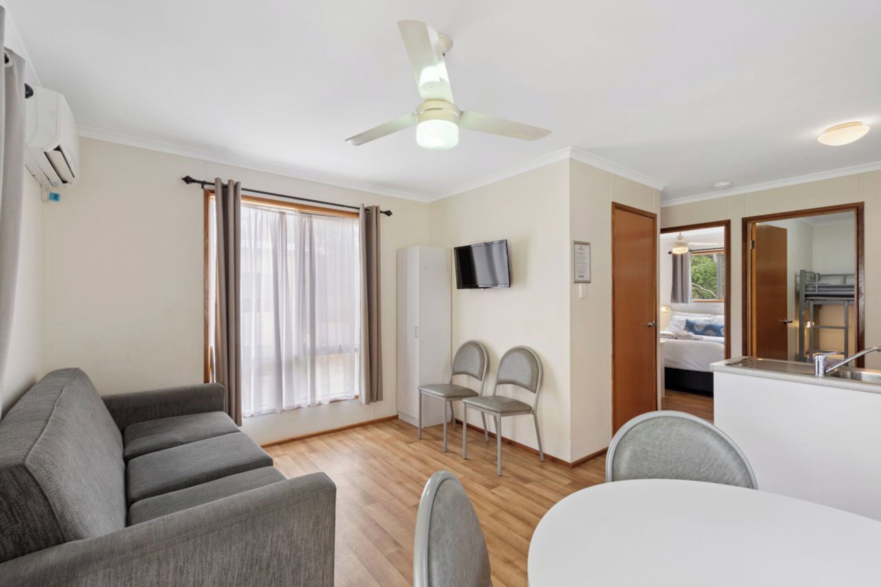 A compact living space with a sofa, two chairs, a small dining table, a ceiling fan, a TV, and a kitchenette.