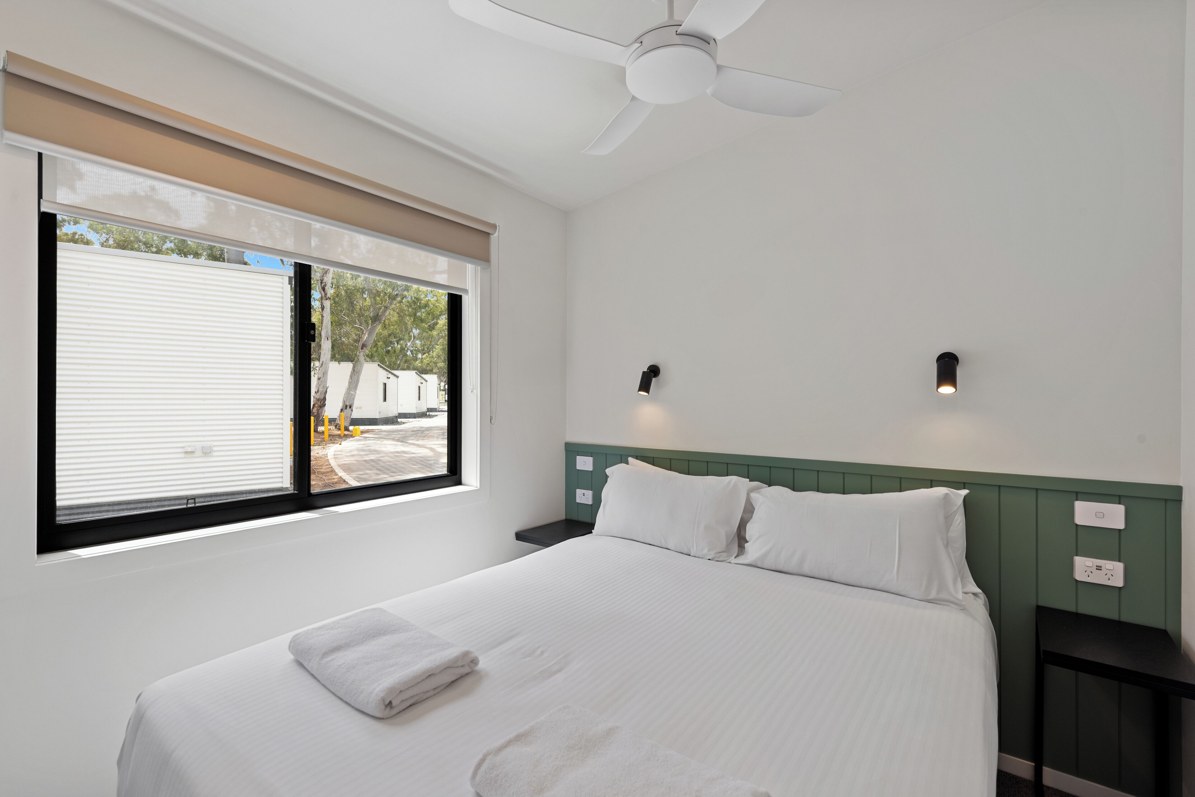 Habitación de hotel con cama, ventilador de techo, ventana con persianas y vistas al exterior.