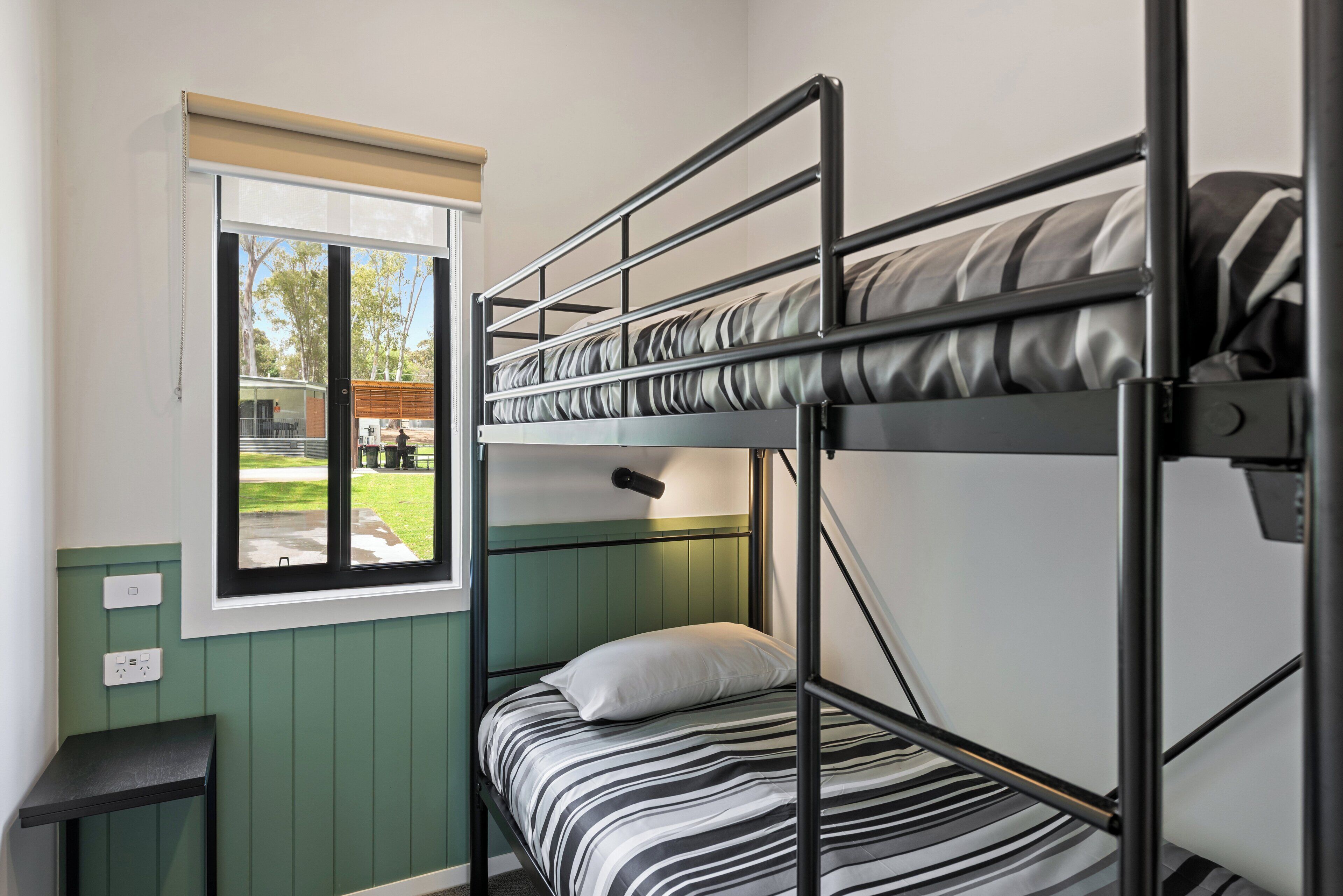 Un lit superposé avec un matelas à rayures, une petite table de chevet, une fenêtre donnant sur l’extérieur et une prise électrique au mur.