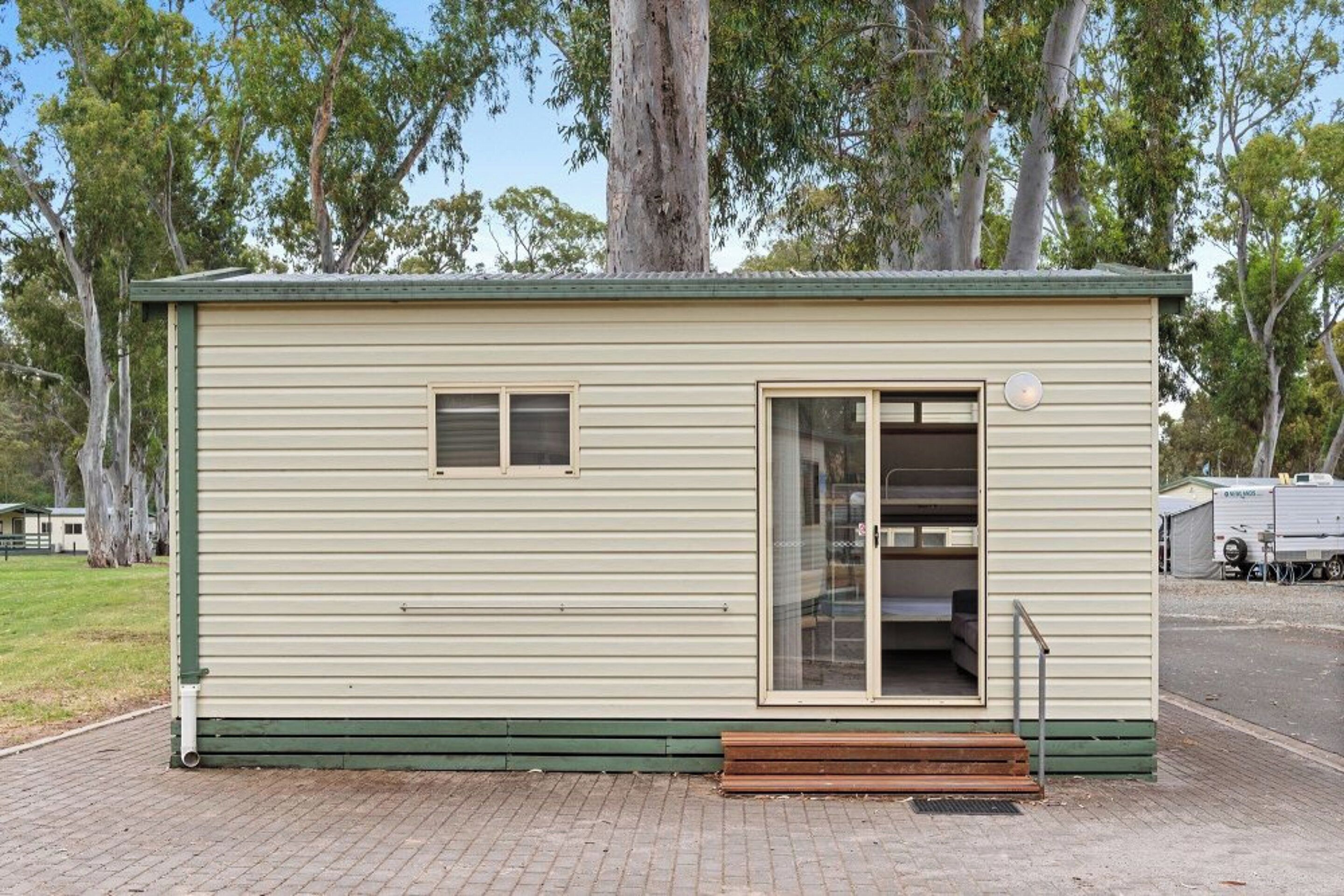 A small, single-story building with a green door and windows, a bench outside, and trees in the background.