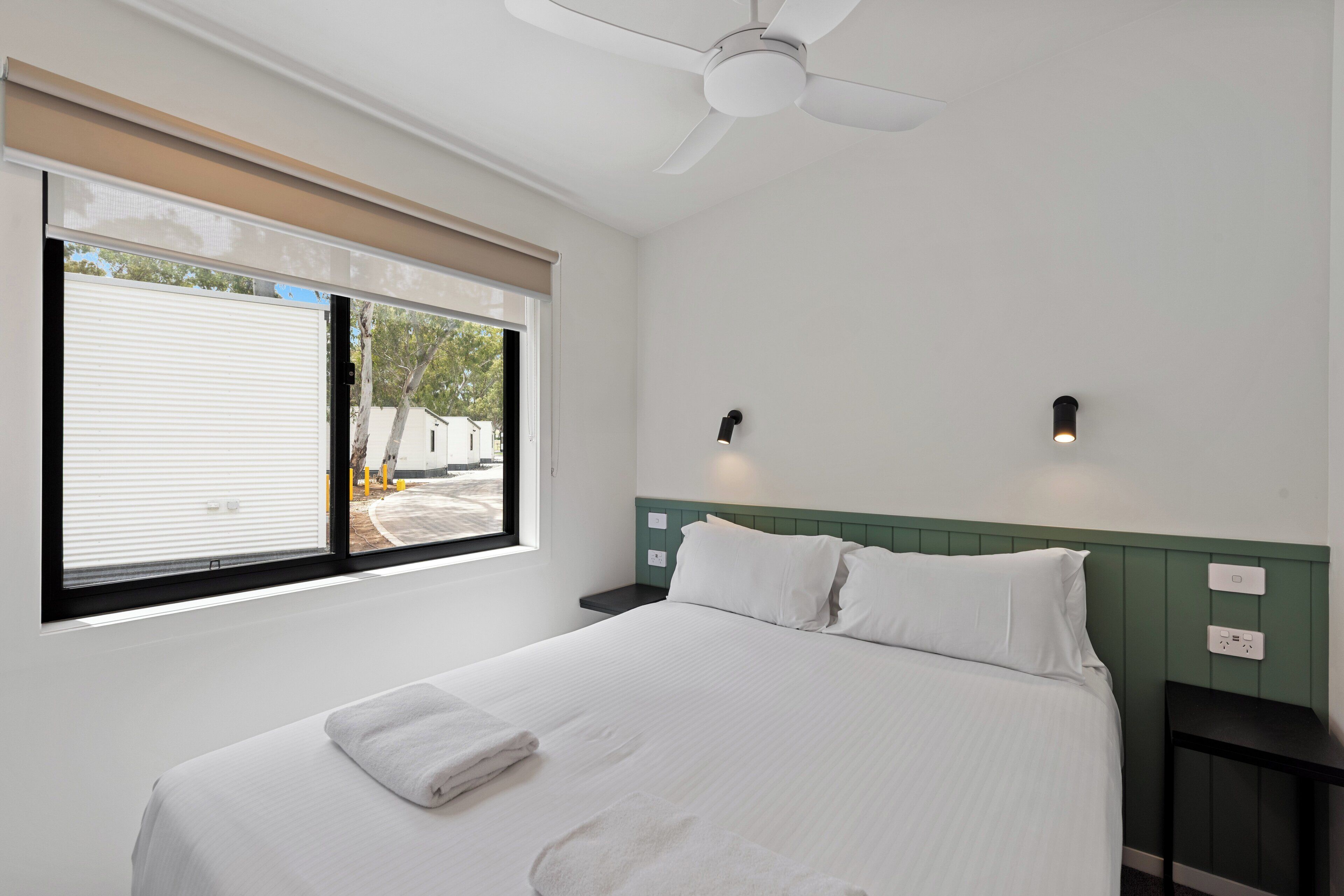 A neatly made bed with white linens and a folded towel, a ceiling fan, and a window with blinds.