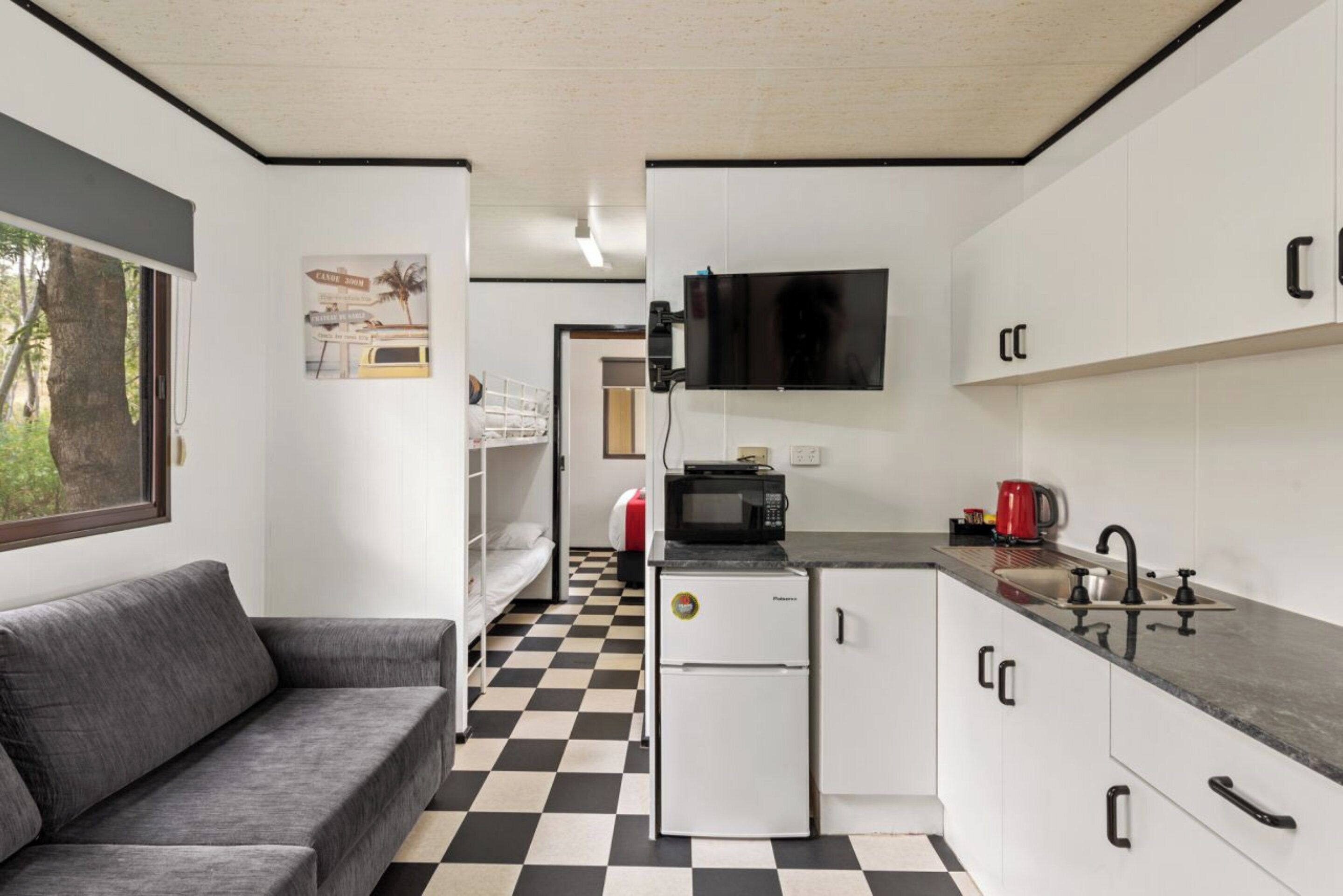 A compact kitchen with a black and white checkered floor, a white refrigerator, a microwave, a sink, and a television mounted above it.