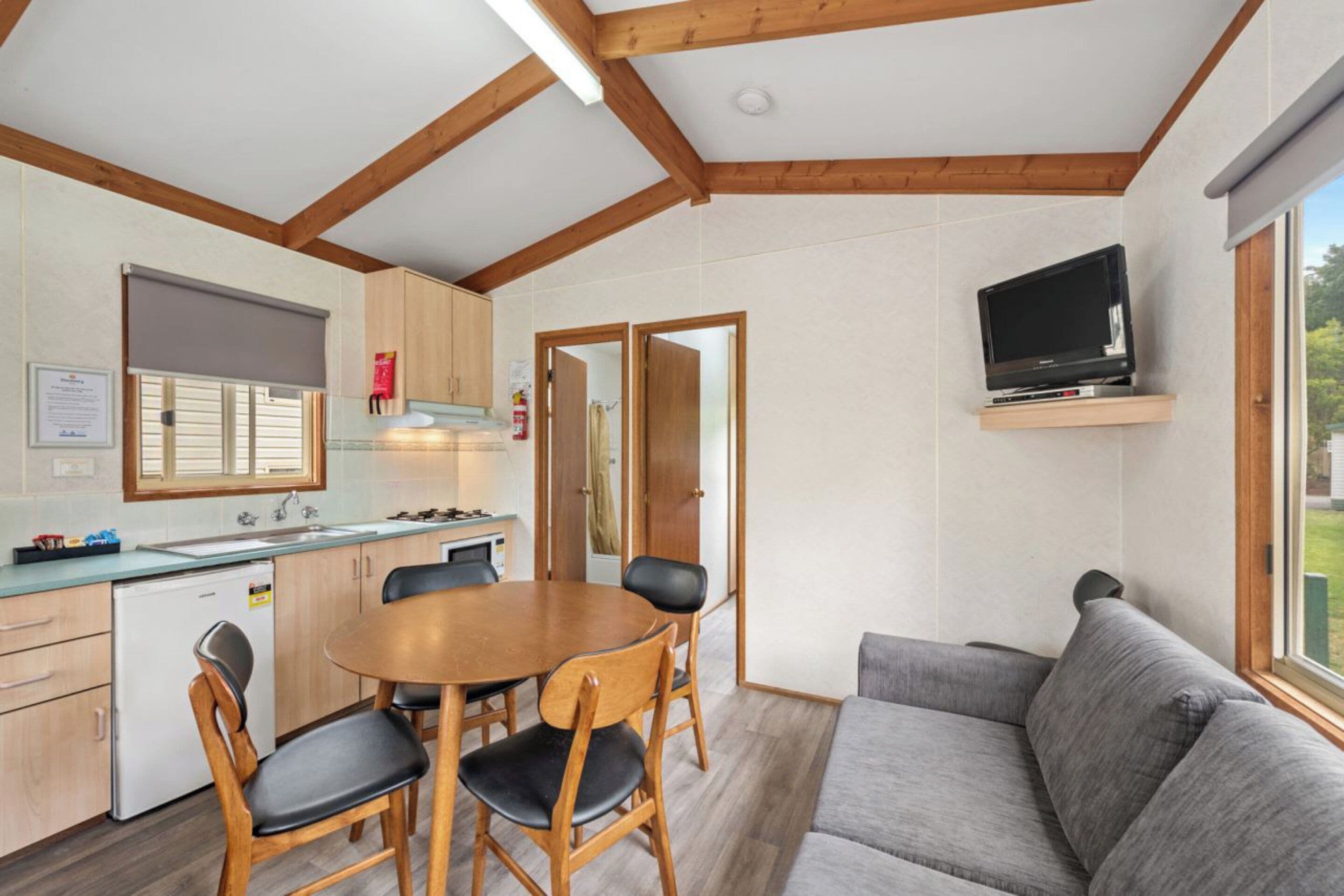 A compact kitchen with a dining area, a grey sofa, and a TV mounted on the wall.