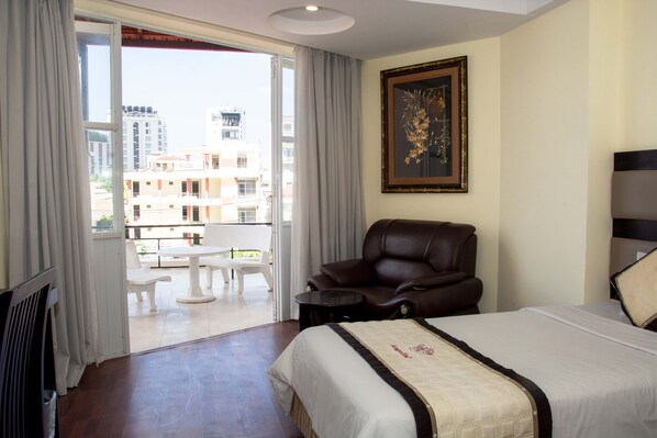 A hotel room with a bed, a leather chair, a small table, and a balcony with a view of buildings. - Angella (Nha Trang)