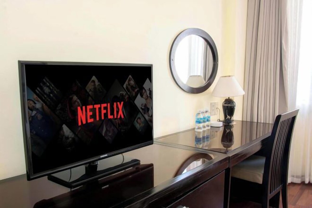 Una camera d'albergo con una TV a schermo piatto che mostra il logo di Netflix, uno specchio rotondo, un tavolino in vetro, una sedia e una lampada.