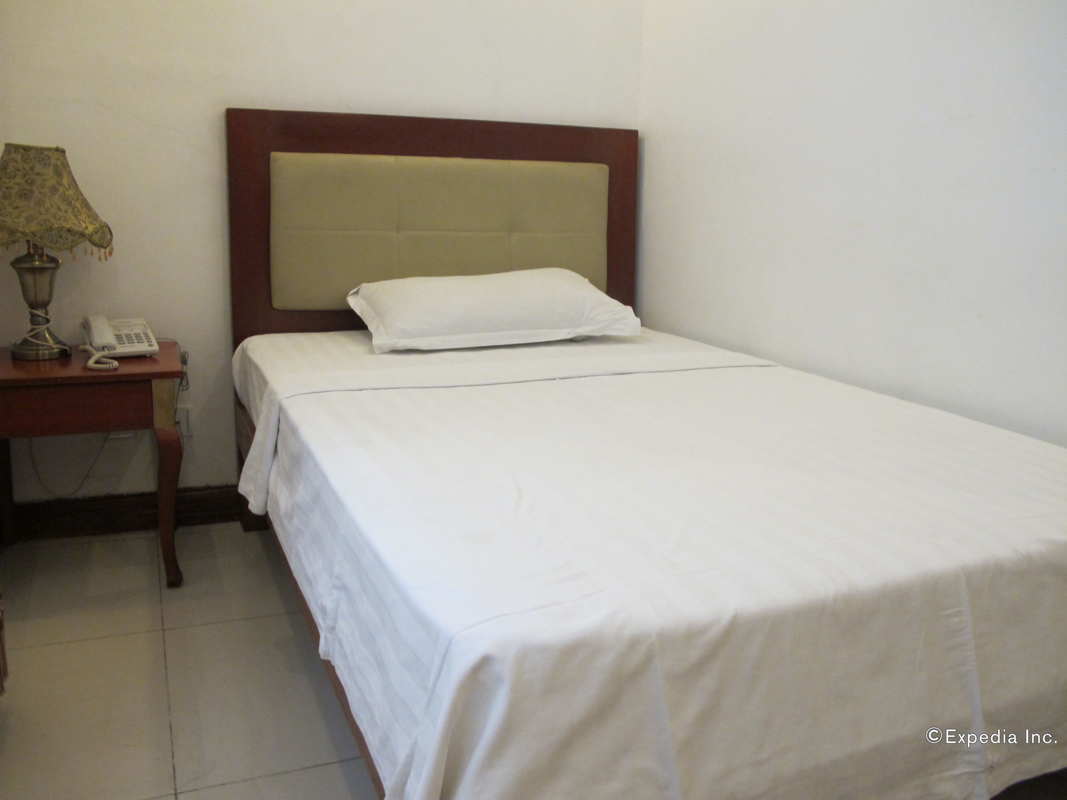 A single bed with a wooden headboard, a bedside table with a lamp and a telephone, and a plain white wall.