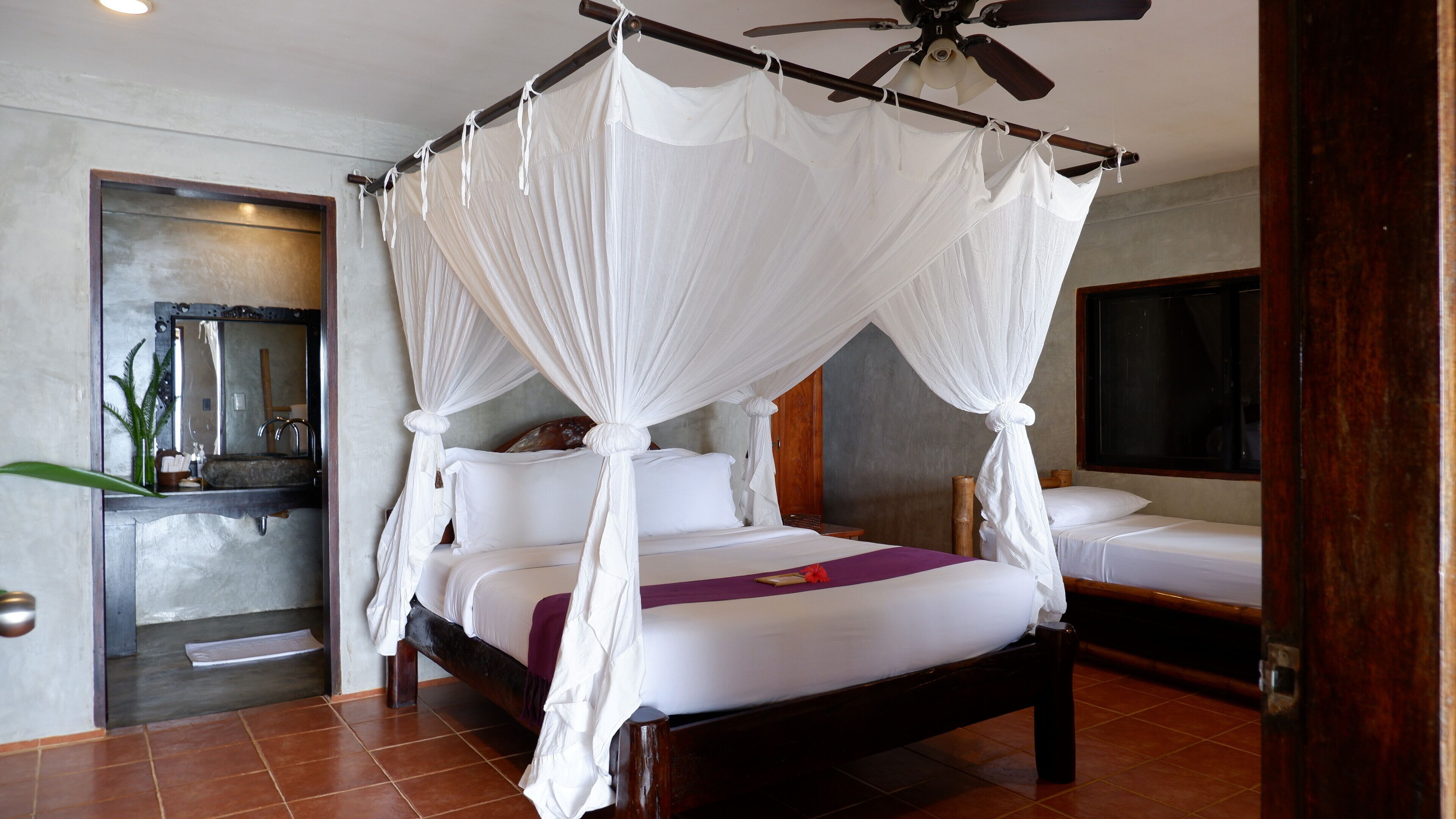 A four-poster canopy bed with a wooden frame and white bedding, a ceiling fan, and a small bedside table with a lamp.