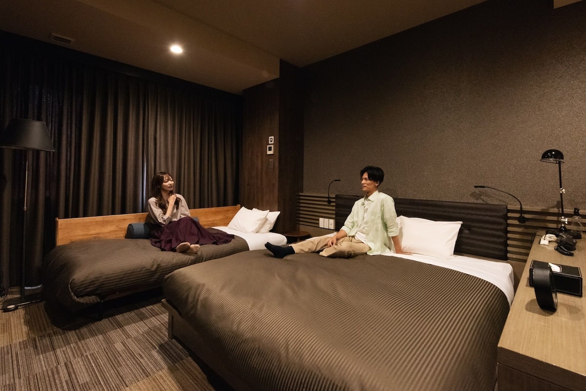 Une chambre d’hôtel avec deux lits, un rideau sombre en arrière-plan, une lampe et une table de chevet.