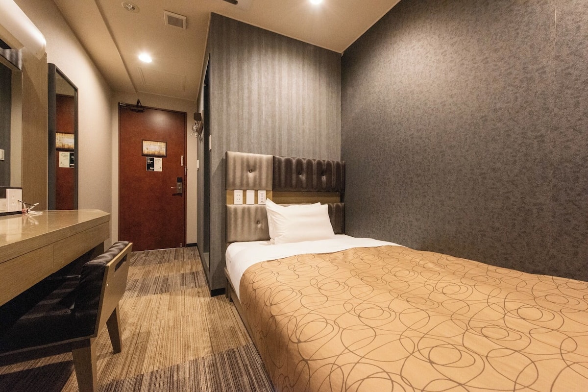 Une chambre d’hôtel avec un lit, un bureau en bois, un miroir et une tête de lit fixée au mur.