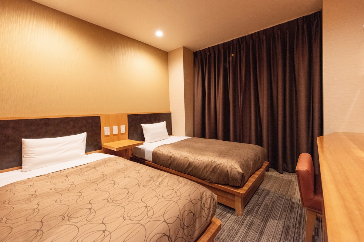 Une chambre d’hôtel avec deux lits, une tête de lit en bois, un bureau et une grande fenêtre avec des rideaux.
