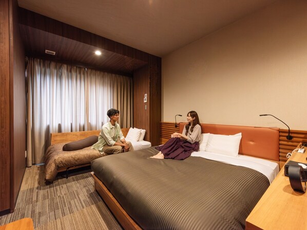 A hotel room with a large bed, a smaller sofa, a desk with a lamp, and a window with curtains.