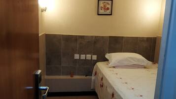 A small, well-lit room with a bed, a wall-mounted light, and a framed picture above the bed.