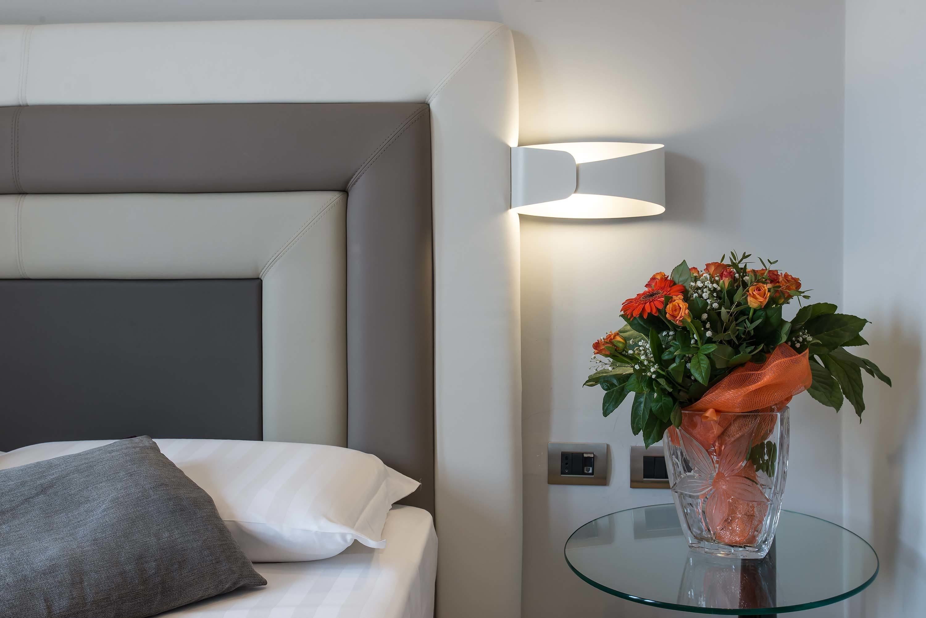 A modern hotel room with a bed, a bedside table with a vase of flowers, and a wall-mounted light fixture.