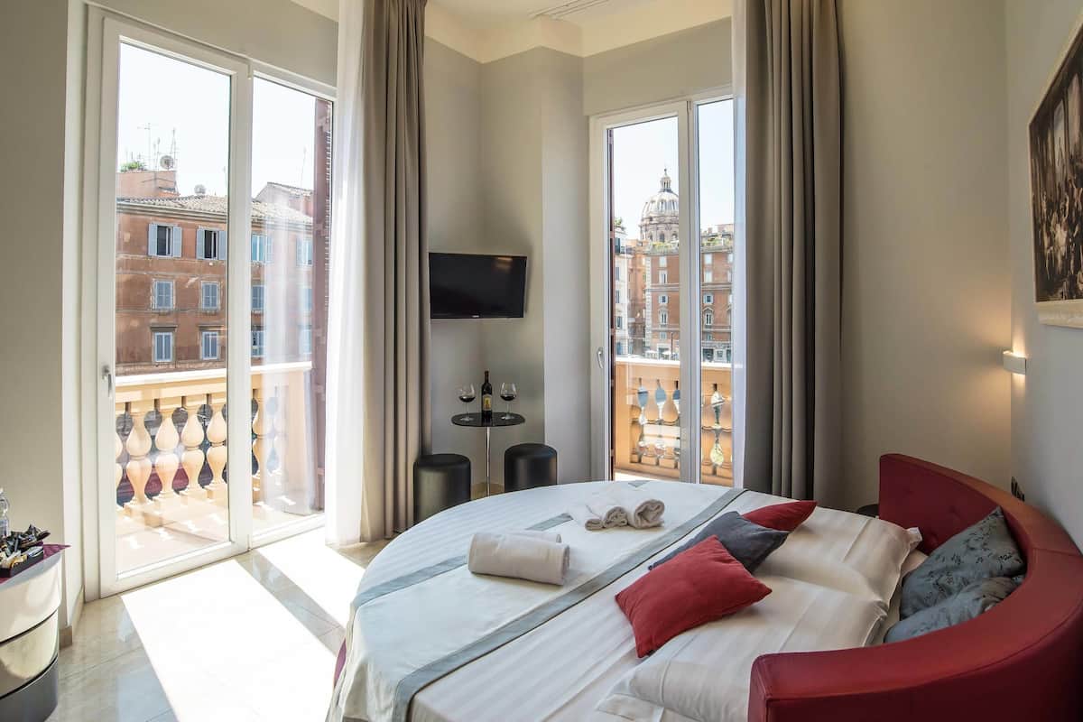 A hotel room with a large bed, a TV mounted on the wall, and a balcony with a view of buildings.