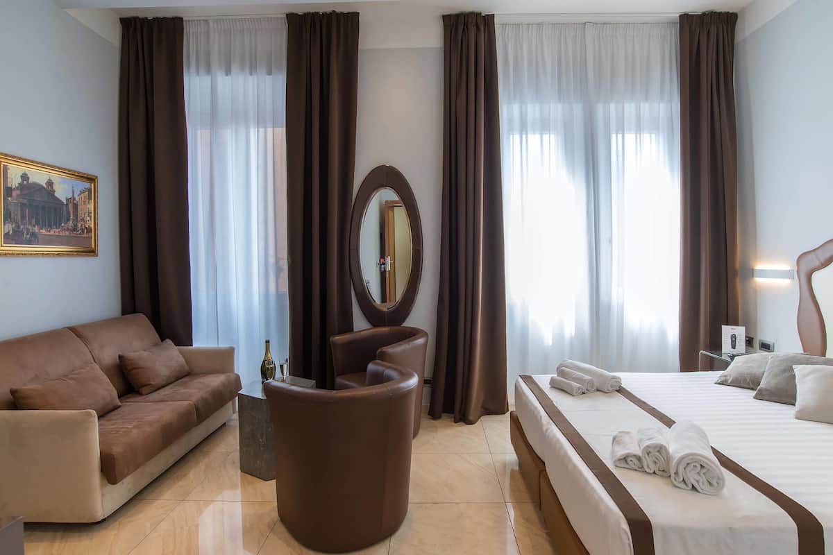 A hotel room with a bed, a sofa, a chair, a round mirror, and a painting on the wall.