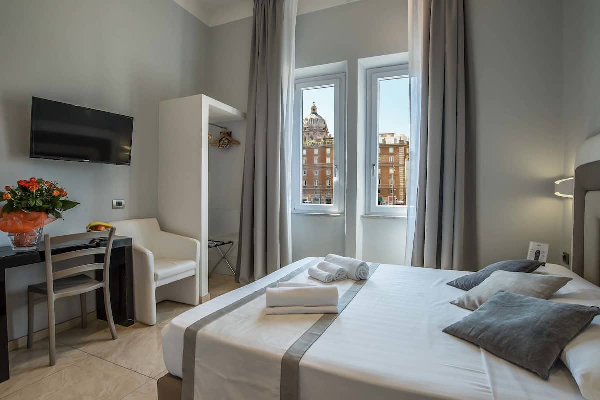 A hotel room with a bed, a desk, a chair, a TV, and a window with a view of buildings.