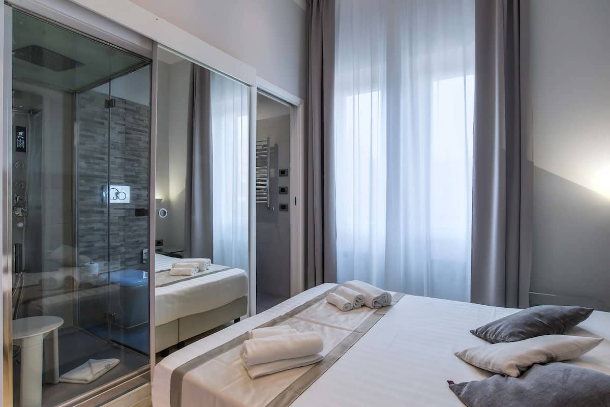 A modern hotel room with a glass-enclosed shower, a bed with neatly folded towels, and a view of the city through large windows.