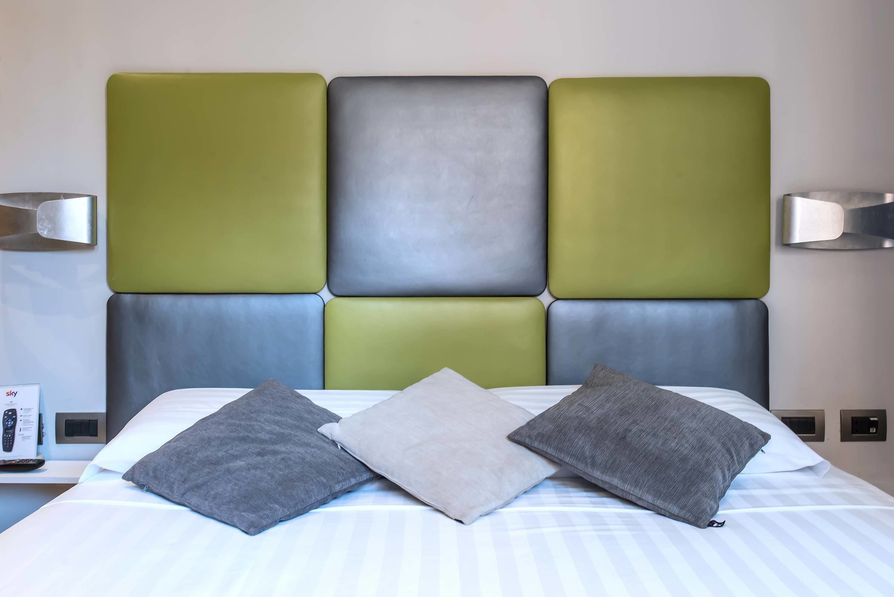 Hotellihuone, jossa on sänky, harmaat ja valkoiset tyynyt, harmaa pääty ja yöpöytä, jossa on kaukosäädin.