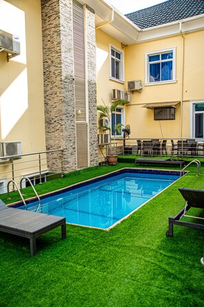 Piscina con césped artificial, sillas de descanso y unidades de aire acondicionado en un día soleado.