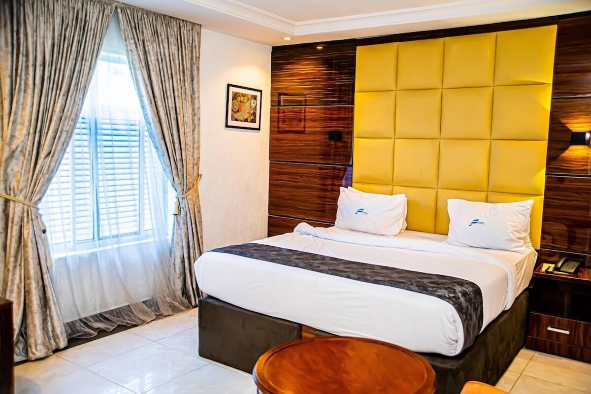Une chambre d’hôtel avec un grand lit, une tête de lit en bois, une table de chevet et une fenêtre avec des rideaux.
