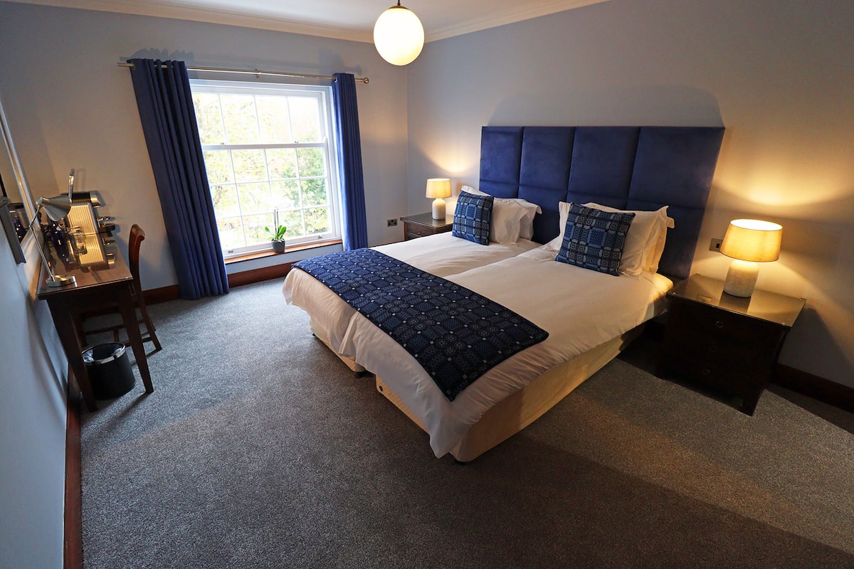 A neatly arranged hotel room with a large bed, bedside lamps, a desk with a chair, and a window with curtains.