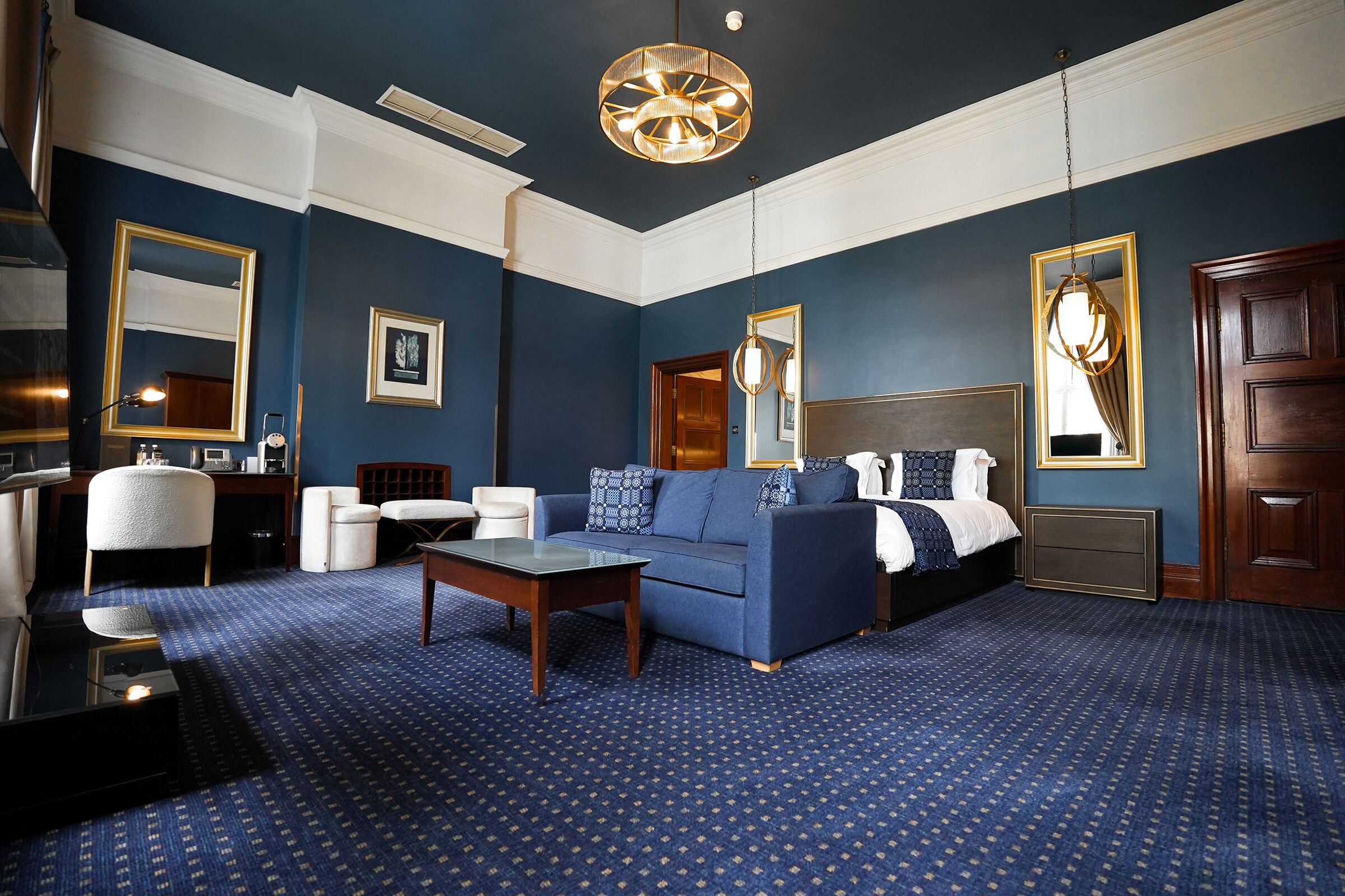 A hotel room with a blue carpet, a sofa, a coffee table, a bed, a wooden door, and a mirror with a gold frame.