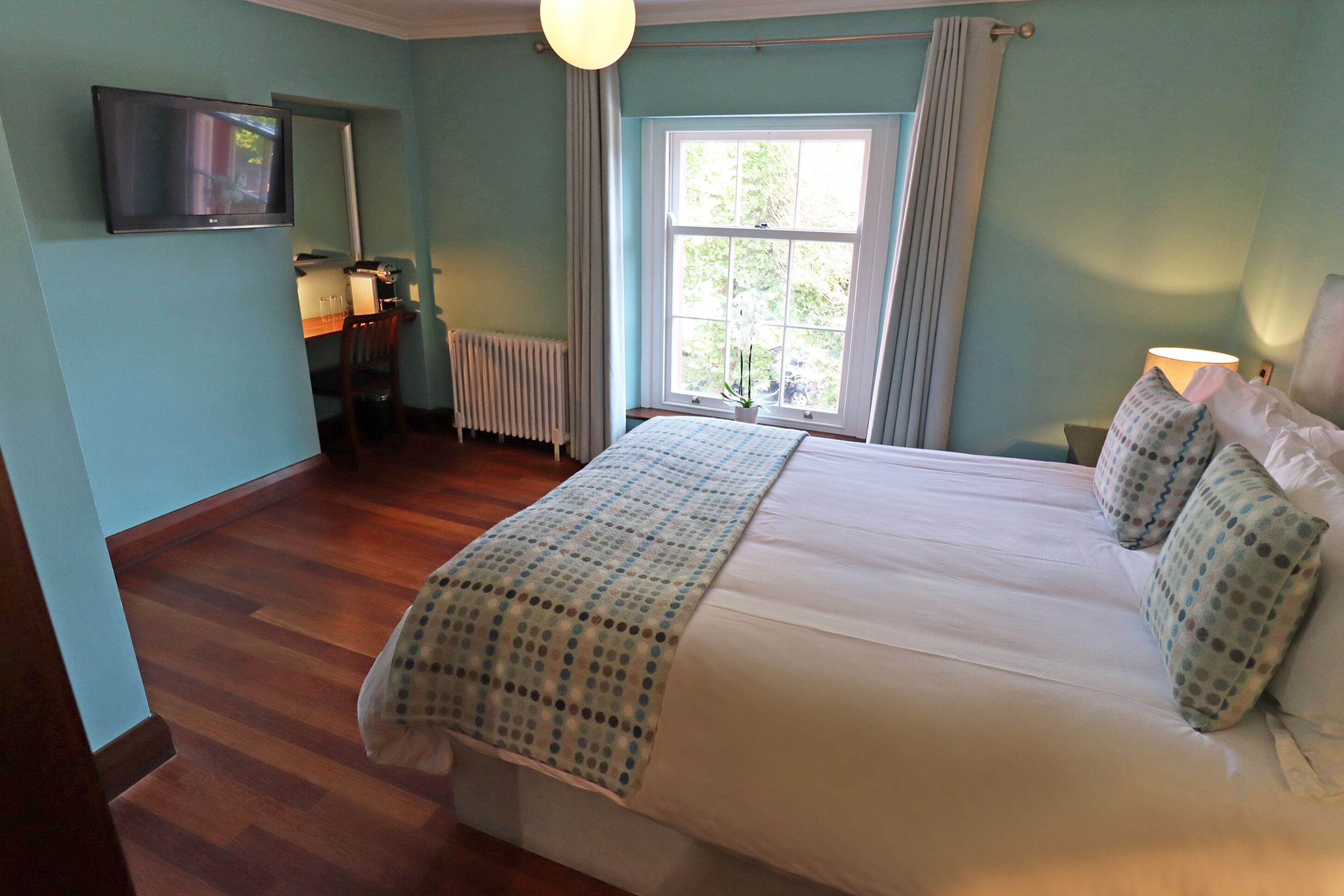 A bedroom with a bed, a desk, a chair, a radiator, a TV, and a window with curtains.