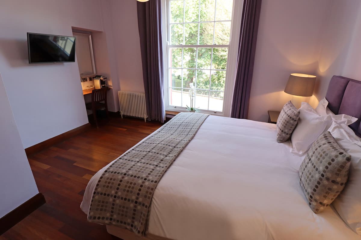 A bedroom with a bed, a TV mounted on the wall, a window with curtains, a bedside lamp, and a radiator.