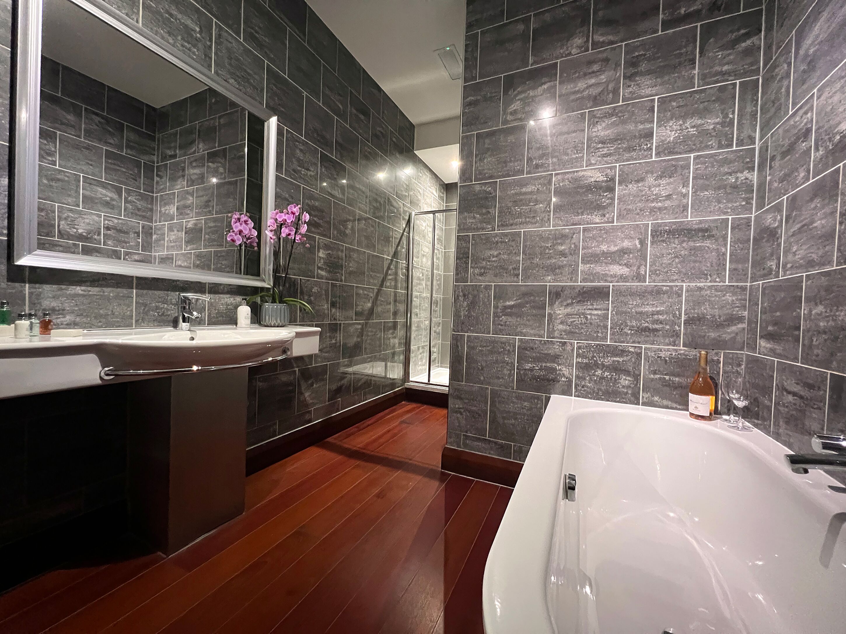 A modern bathroom with a large mirror, a white bathtub, and dark tiled walls.