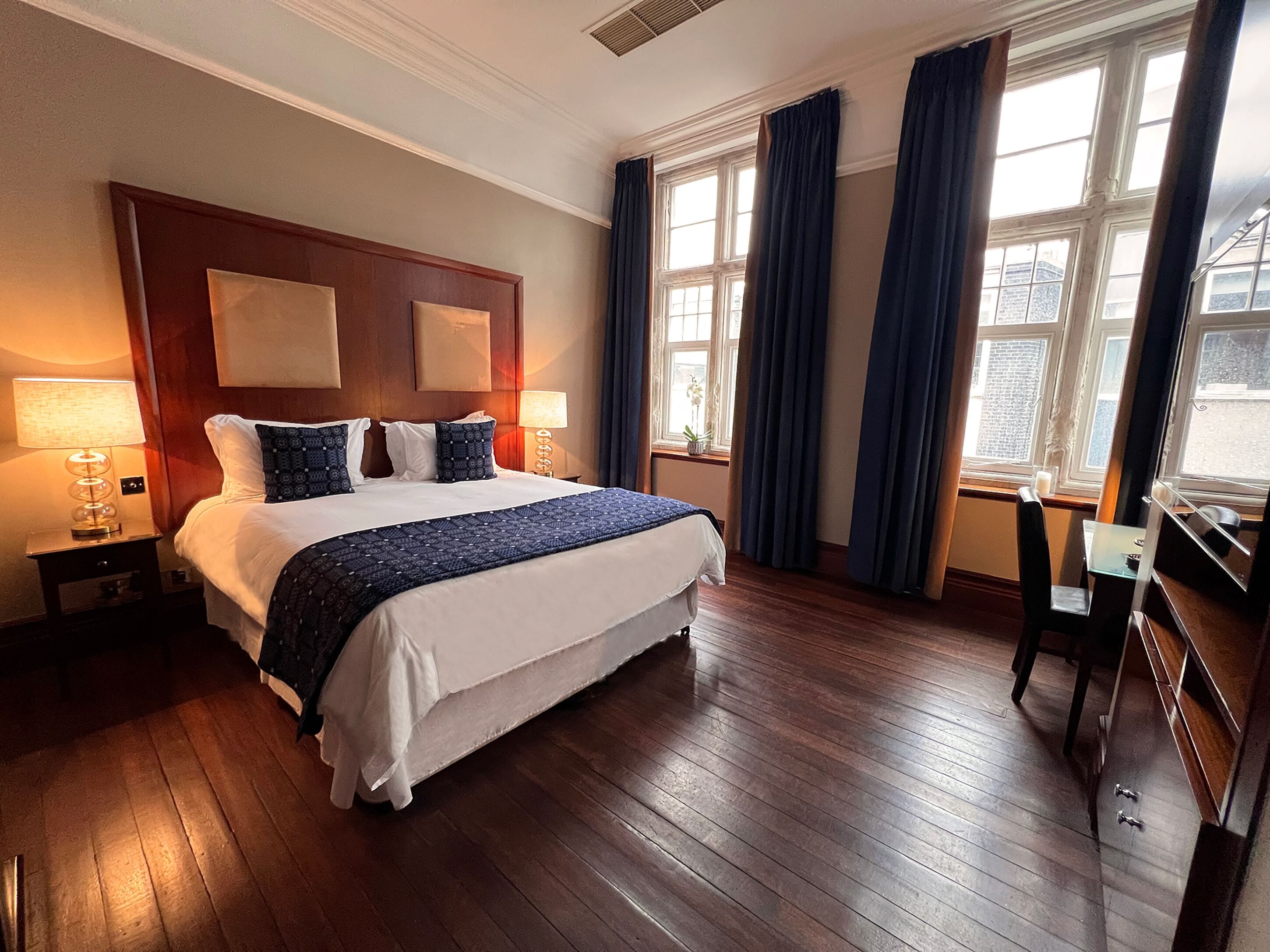 A hotel room with a large bed, wooden flooring, a desk, and a window with curtains.