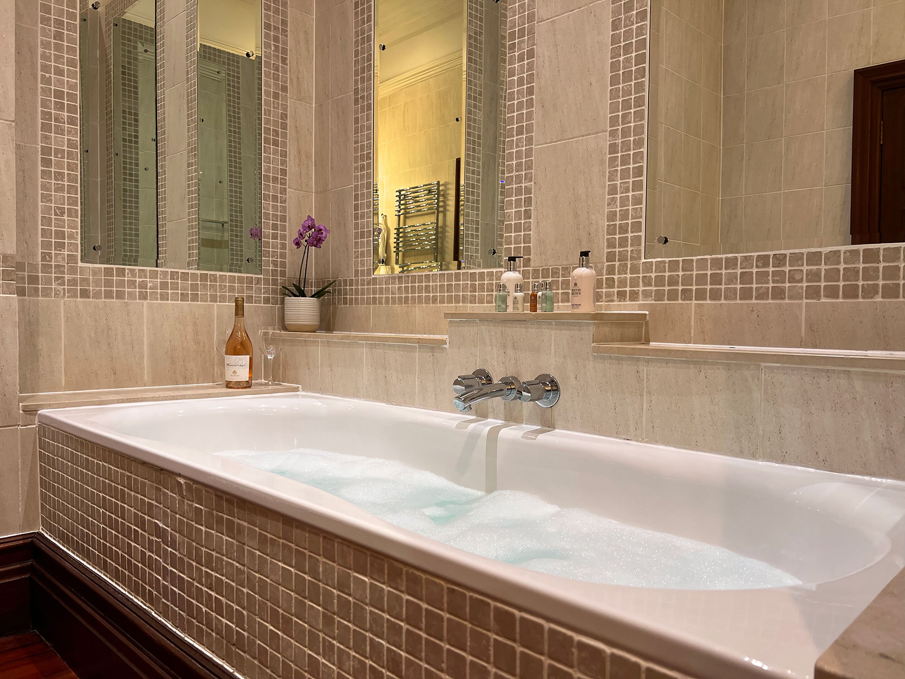 A large bathtub with a glass door, surrounded by beige-tiled walls and a mirror.
