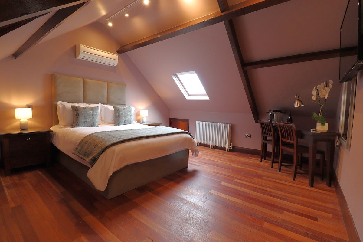 A bedroom with a large bed, a skylight, wooden floors, a desk with chairs, and a radiator.