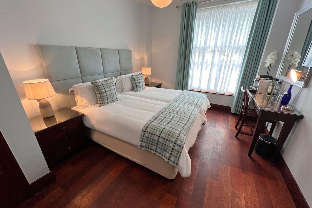 A hotel room with a large bed, two bedside tables with lamps, a wooden floor, a window with curtains, and a small table with a vase.